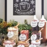 jars of toppings for Hot Chocolate Bar. Toppings are labeled on each jar and include mini marshmallows, peppermint sticks, chocolate chips, peppermint chips, toffee bits, and kosher salt. Bottles of chocolate sauce and caramel sauce sitting behind. Sign hanging behind says "Hot Chocolate Bar warm up buttercup"