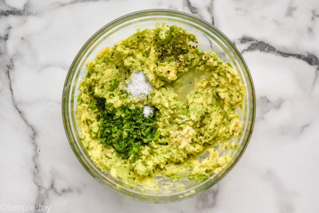 Overhead view of glass bowl of avocado and seasonings for Seven layer dip recipe.