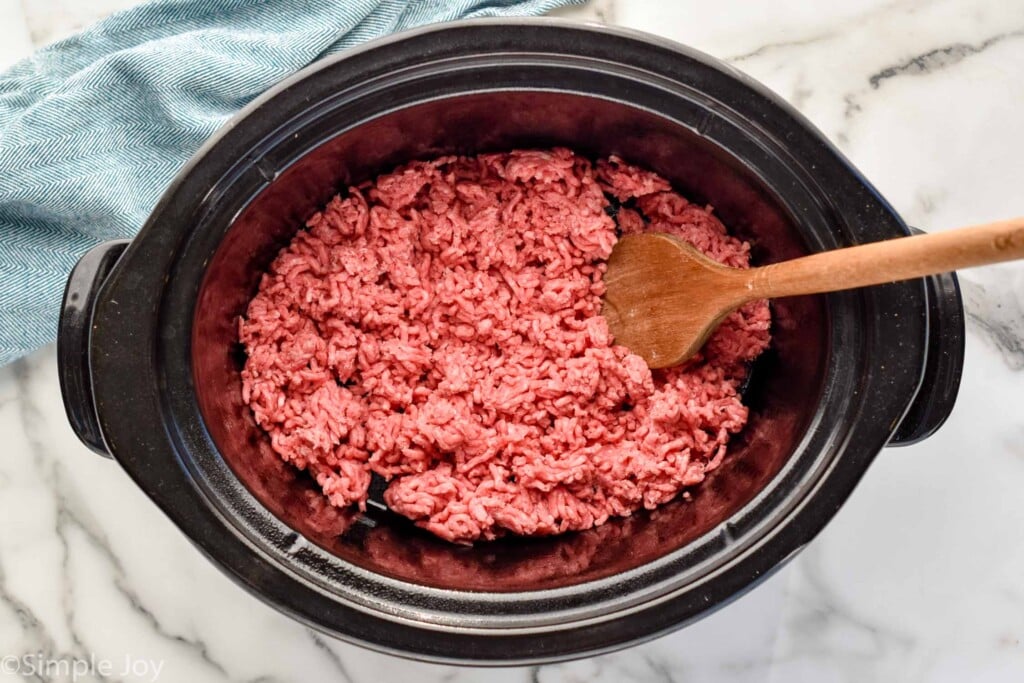 Overhead view a crock pot with hamburger and a wooden spoon for Slow Cooker Taco Meat recipe