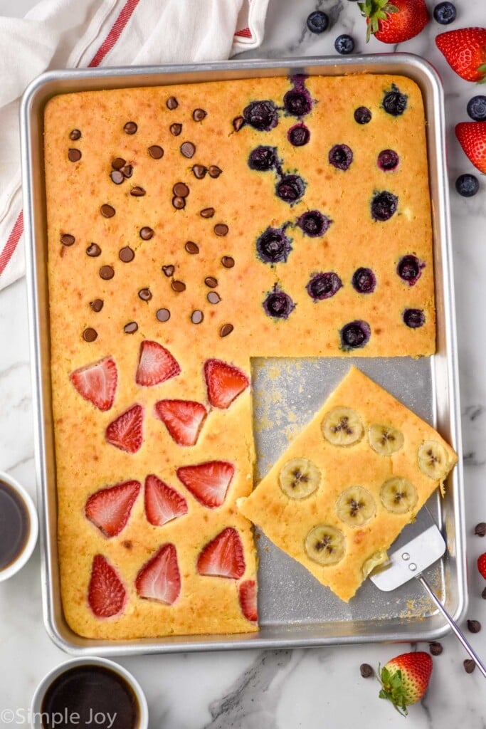 Overhead view of Sheet Pan Pancakes garnished with chocolate chips, blueberries, strawberries, and bananas. Banana square cut out and sitting on serving spatula on baking sheet. Coffee and berries beside.