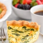 Pinterest graphic for Spinach Quiche recipe. Text says, the best Spinach Quiche simplejoy.com." Image shows a slice of Spinach Quiche on a plate with a fork. Bowl of berries and cup of coffee beside.