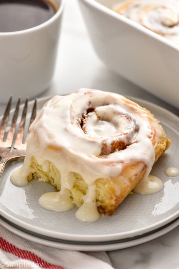 homemade cinnamon roll topped with icing on a plate with a fork sitting beside. Baking dish of cinnamon rolls and mug of coffee sitting in background