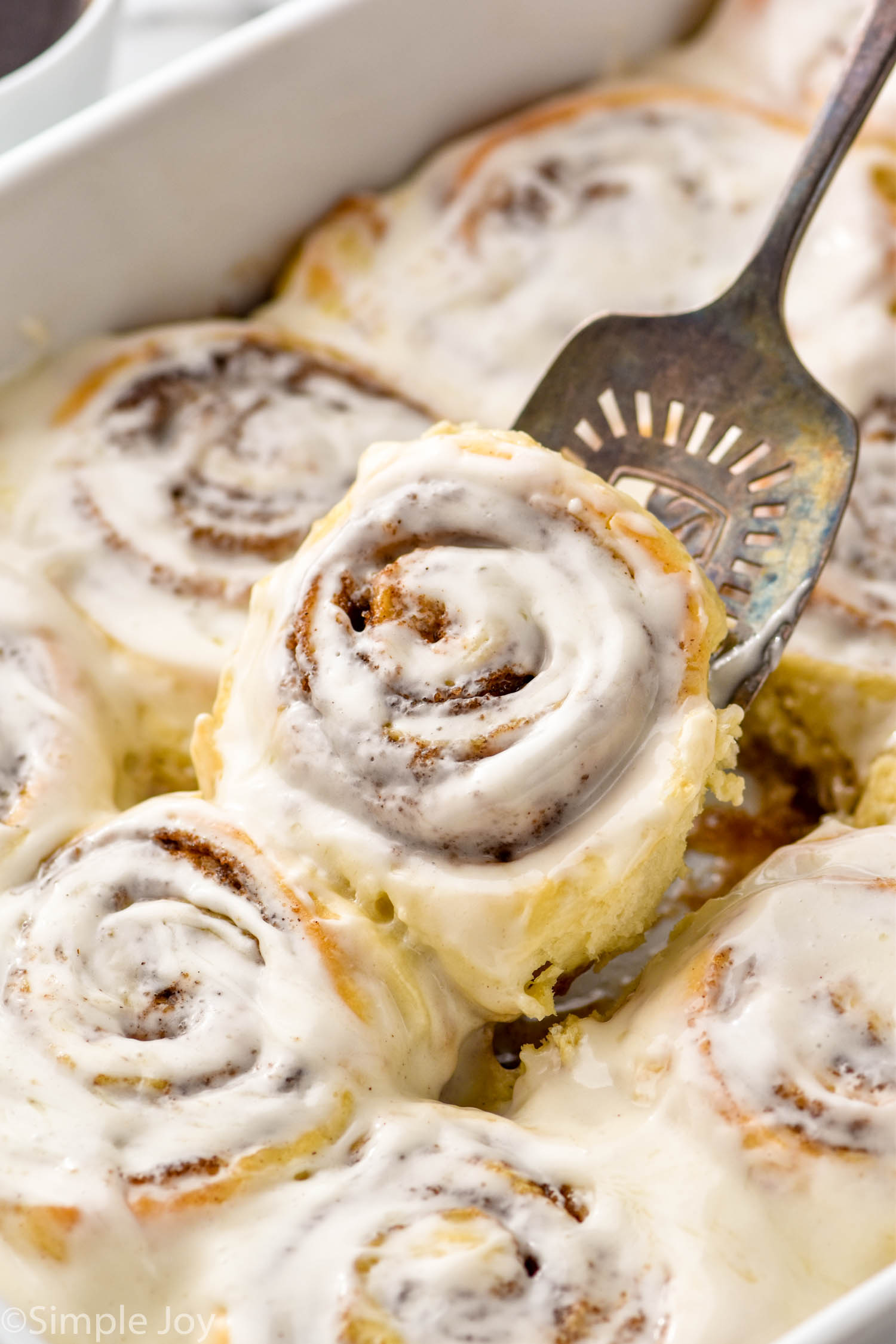 Spatula lifting a homemade cinnamon roll out of baking dish of cinnamon rolls with icing.