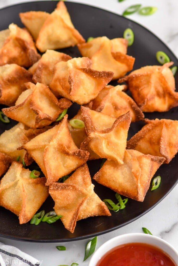 Plate of Crab Rangoon garnished with sliced green onions, bowl of red sauce beside.