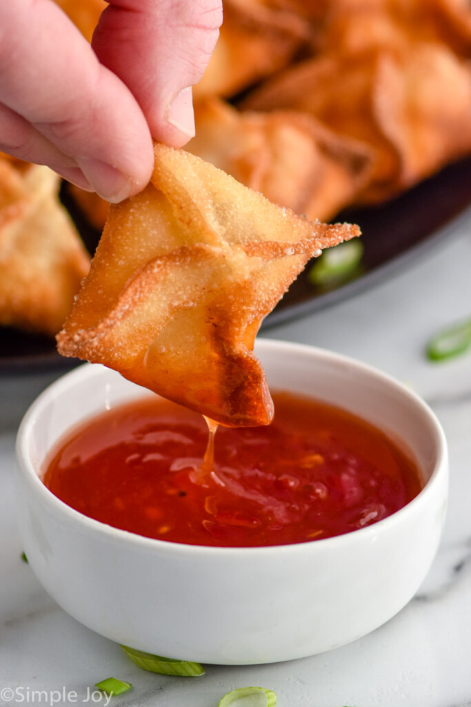 Side view of person's hand dipping Crab Rangoon into small bowl of red sauce. Plate of Crab Rangoon beside.