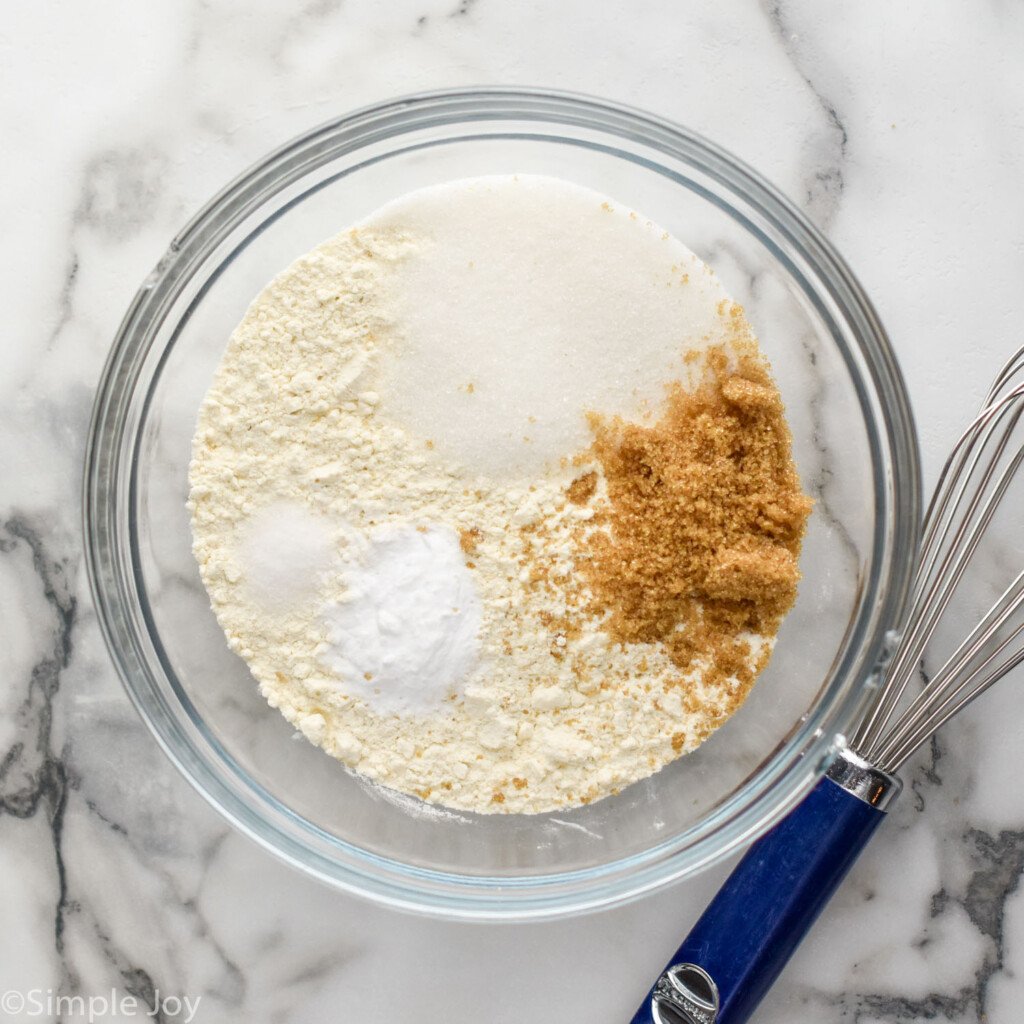 Overhead view of glass mixing bowl of dry ingredients for Sheet Pan Pancakes recipe with whisk beside.