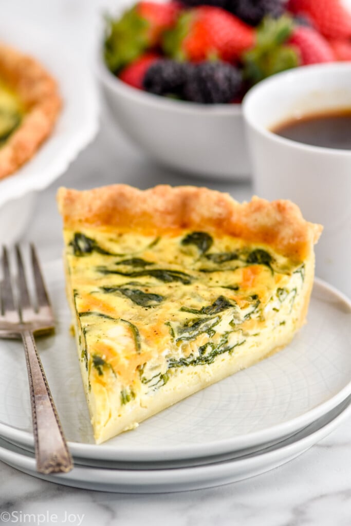 Slice of Spinach Quiche served on a plate with a fork. Cup of coffee and bowl of berries beside.