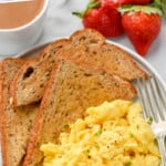 Pinterest graphic for Scrambled Eggs. Says "how to make fluffy scrambled eggs simplejoy.com" Image shows overhead of a plate of Scrambled Eggs, pieces of toast, and a fork. Fresh strawberries sitting beside