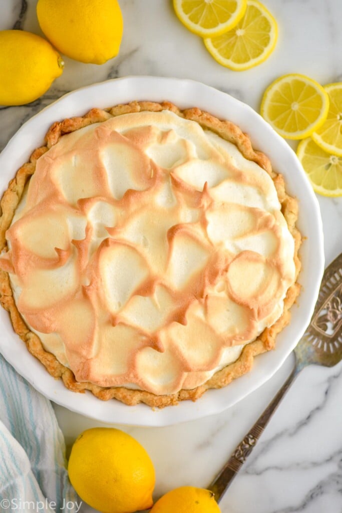 overhead of Lemon Meringue Pie with lemons, lemon slices, and spatula surrounding.