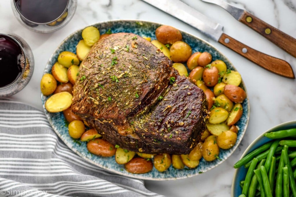Overhead view of a platter with Sirloin Tip Roast and cooked potatoes. Glasses of red wine, carving knives, and bowl of green beans beside.