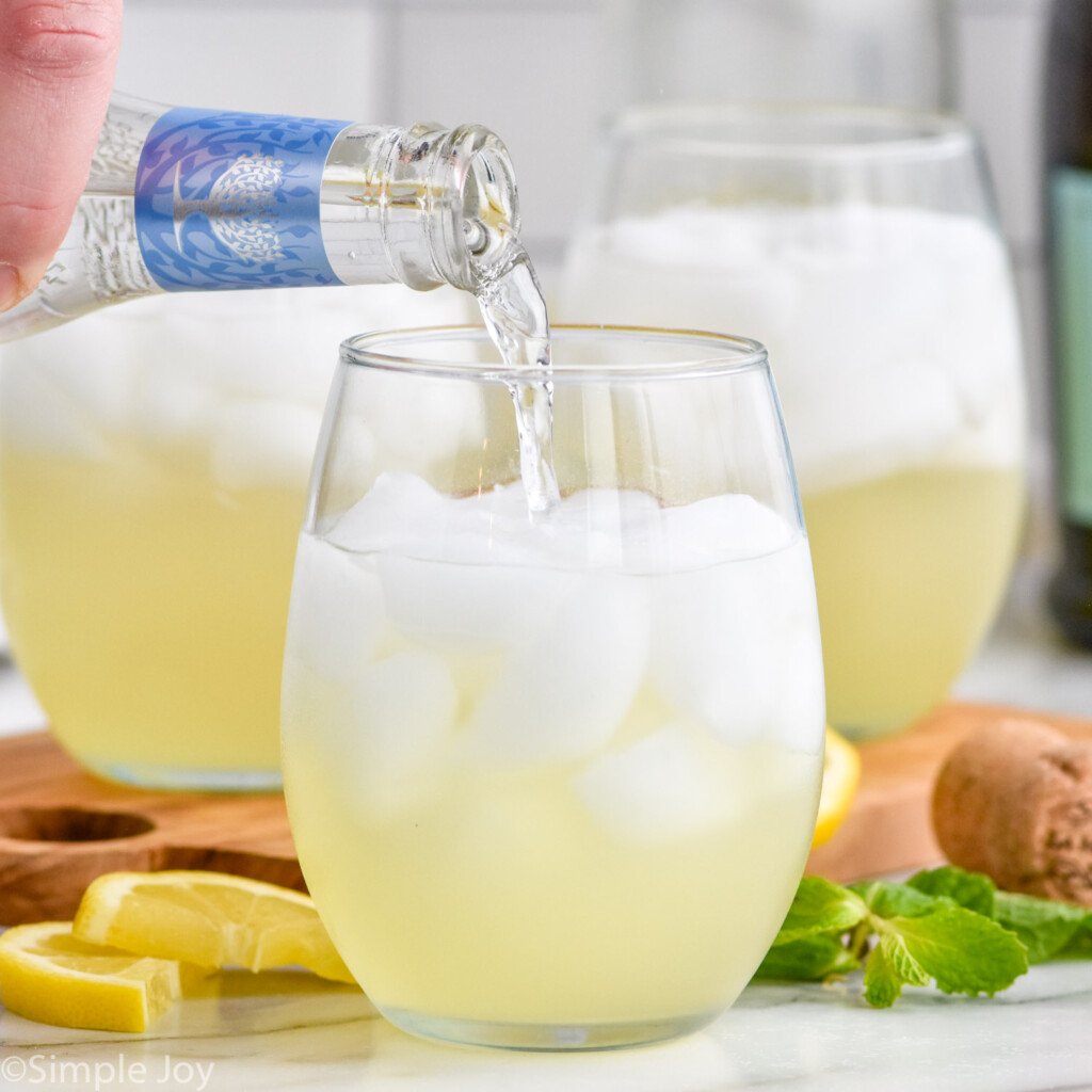 Side view of person's hand pouring bottle of club soda into a glass of ingredients for Limoncello Spritz recipe. Lemon slices and fresh mint leaves for garnish on counter beside.
