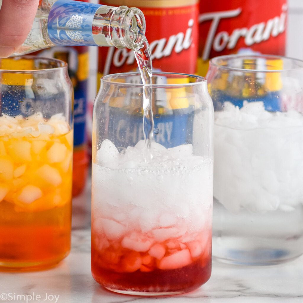 Bottle of club soda pouring into a glass of ice and flavored syrup to make Italian Soda. Two glasses of Italian Soda ingredients and bottles of flavored syrup sitting behind