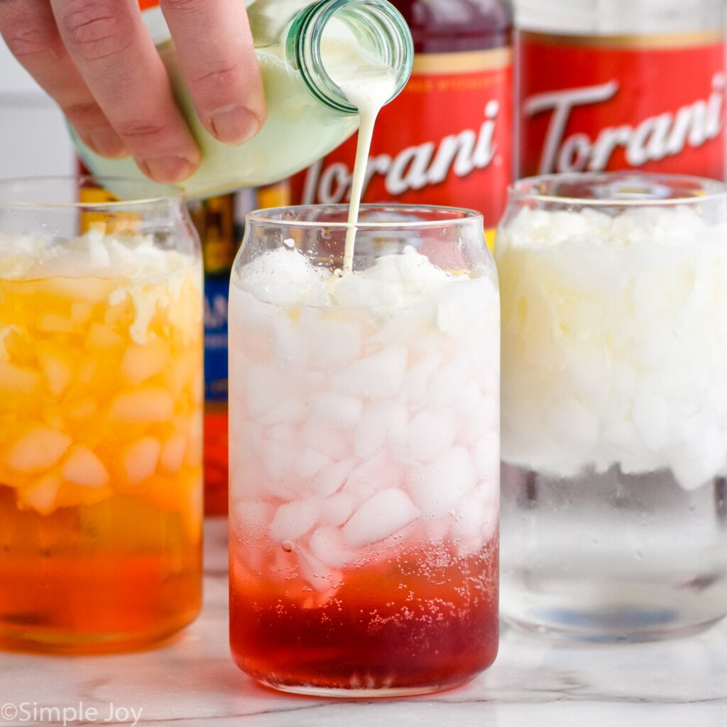 person's hand pouring jar of heavy cream into a glass of ice and Italian Soda ingredients to make an Italian Soda. Two glasses of Italian Soda ingredients and bottles of flavored syrups sitting behind.