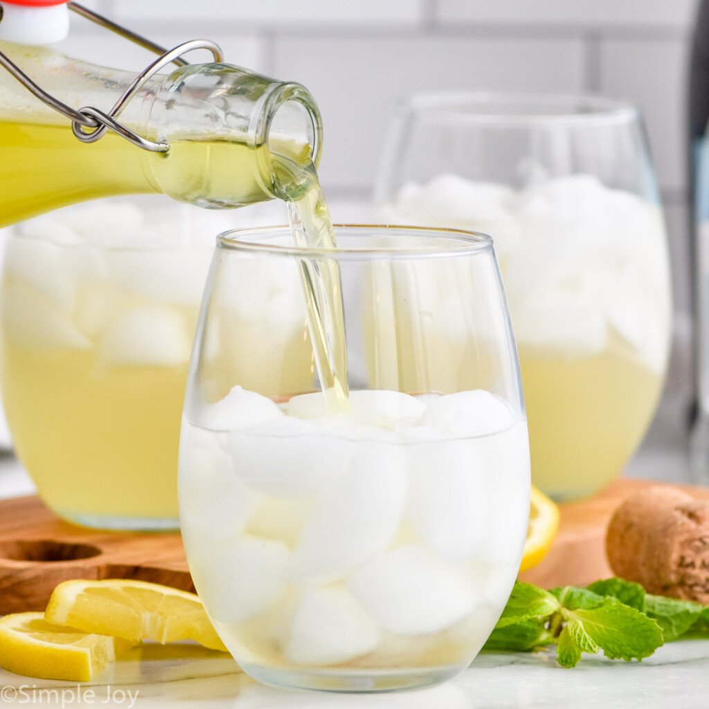 Side view of bottle of limoncello being poured into glass of ingredients for Limoncello Spritz recipe. Lemon slices and mint leaves on counter beside for garnish.