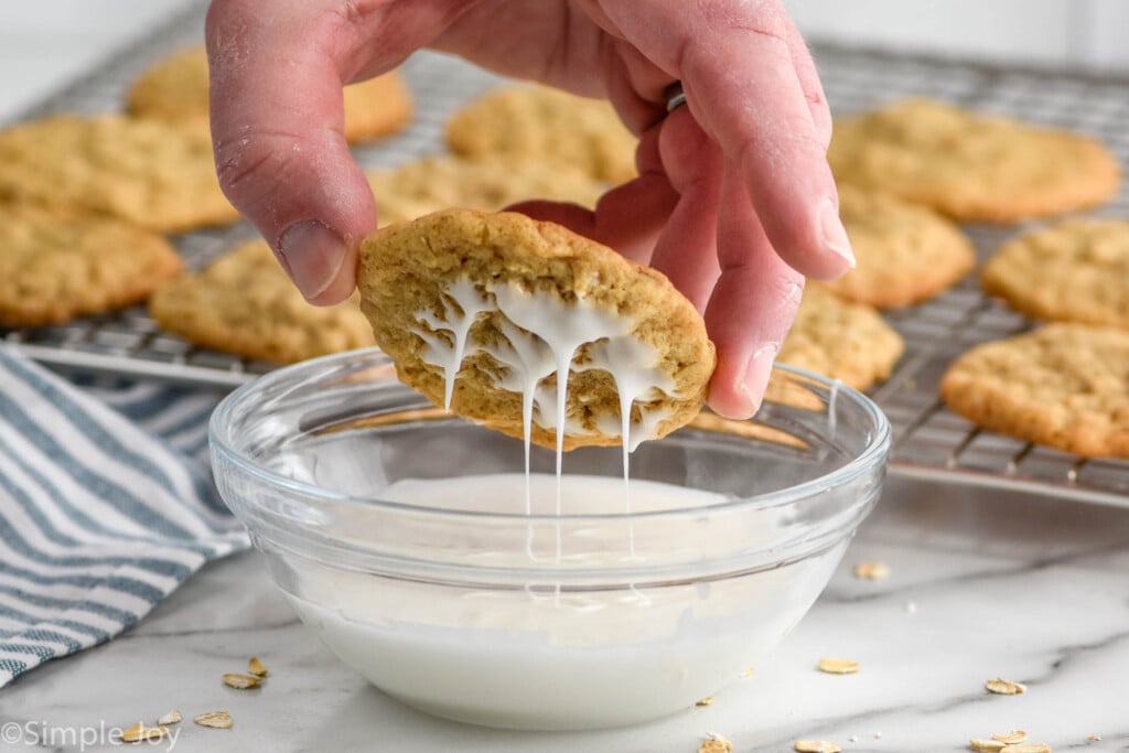 man's hand holding an oatmeal cookie over a bowl of icing, icing dripping off of cookie. Oatmeal cookies on a cooling rack sitting in the background