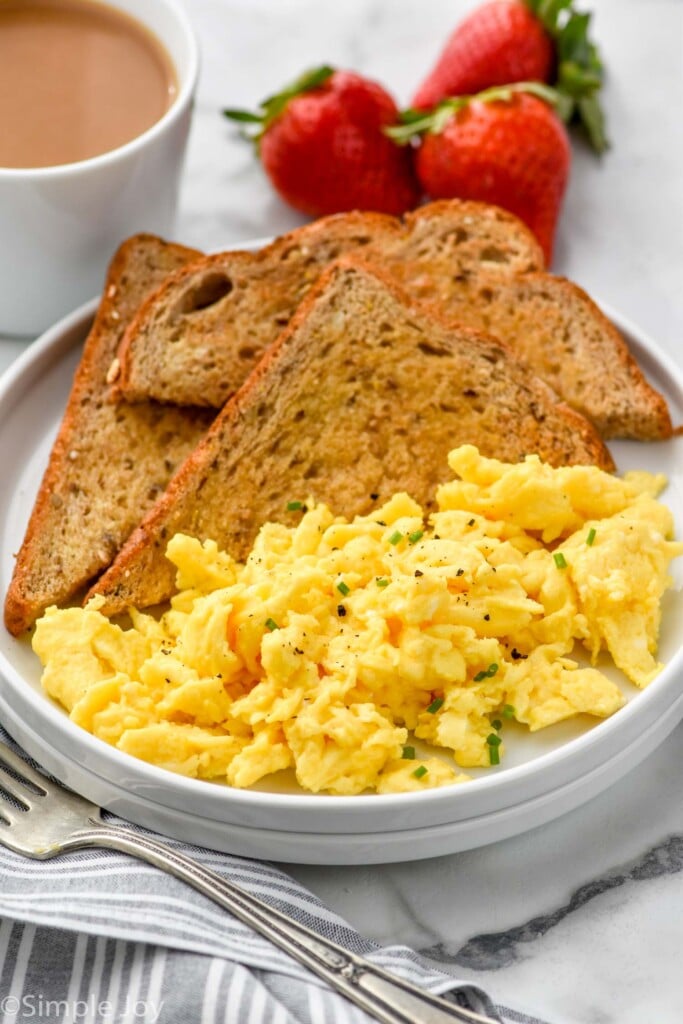 plate of Scrambled Eggs and pieces of toast. Fork, mug of coffee, and fresh strawberries surrounding