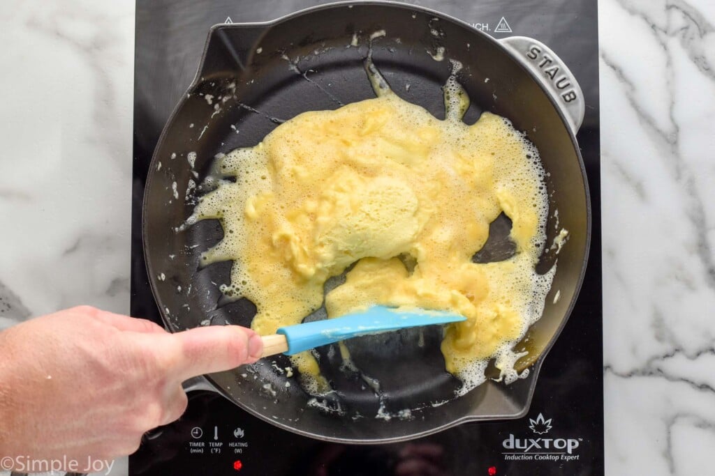 overhead of skillet, man's hand holding spatula and flipping eggs to make Scrambled Eggs