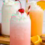 Glass of Italian Soda topped with whipped cream and a cherry with a straw. Two glasses of Italian Soda topped with whipped cream, one with an orange slice, sitting behind. - 10