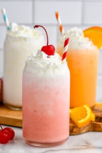 Glass of Italian Soda topped with whipped cream and a cherry with a straw. Two glasses of Italian Soda topped with whipped cream, one with an orange slice, sitting behind.