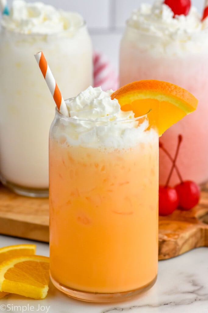 Glass of Italian Soda with a straw, topped with whipped cream and an orange slice. Two glasses of Italian Soda topped with whipped cream sitting behind.