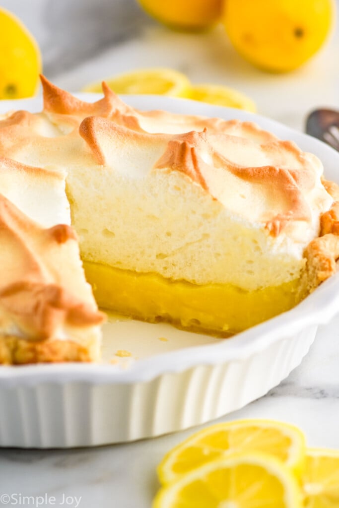 pie plate of Lemon Meringue Pie with slice missing, lemons and lemon slices sitting in front and behind