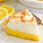 piece of Lemon Meringue Pie on a plate with fork. Lemon, lemon slices, and Lemon Meringue Pie sitting in background.