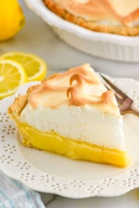 piece of Lemon Meringue Pie on a plate with fork. Lemon, lemon slices, and Lemon Meringue Pie sitting in background.