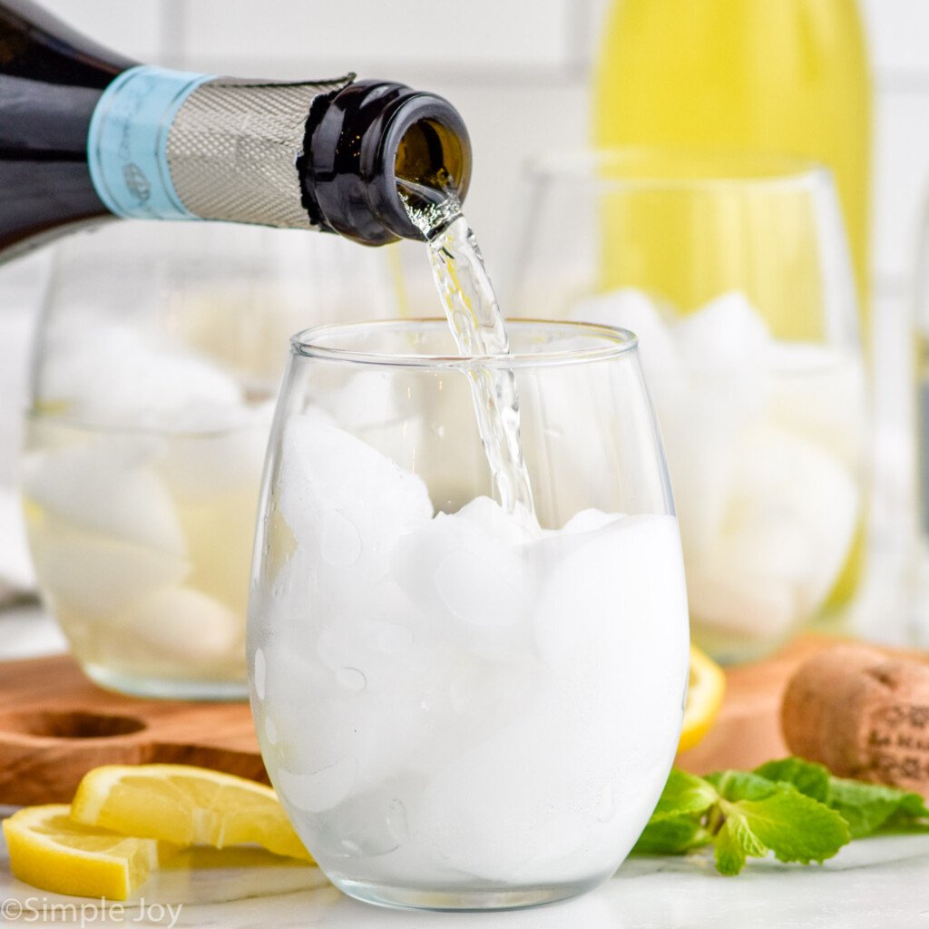 Side view of person's hand pouring bottle of prosecco in to a glass of ice for Limoncello Spritz recipe. Lemon wedges, cork, and mint leaves on counter beside.