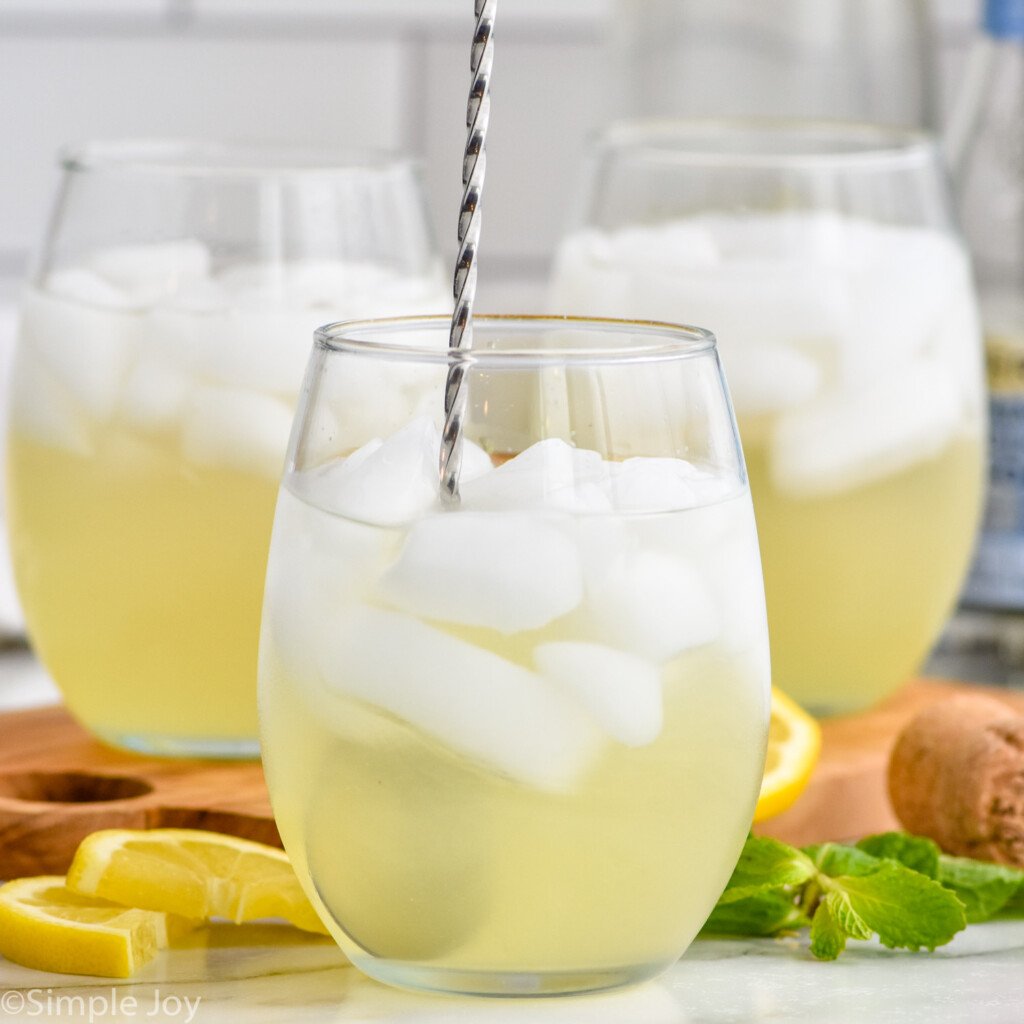 Side view of Limoncello Spritz with ice and a stir stick. Lemon wedges, mint leaves, and a cork beside.