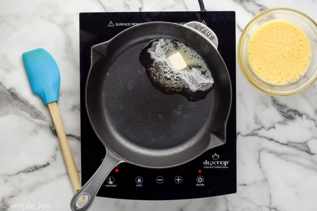 overhead of skillet on a cooktop with butter melting to make Scrambled Eggs. Spatula and bowl of eggs sitting beside