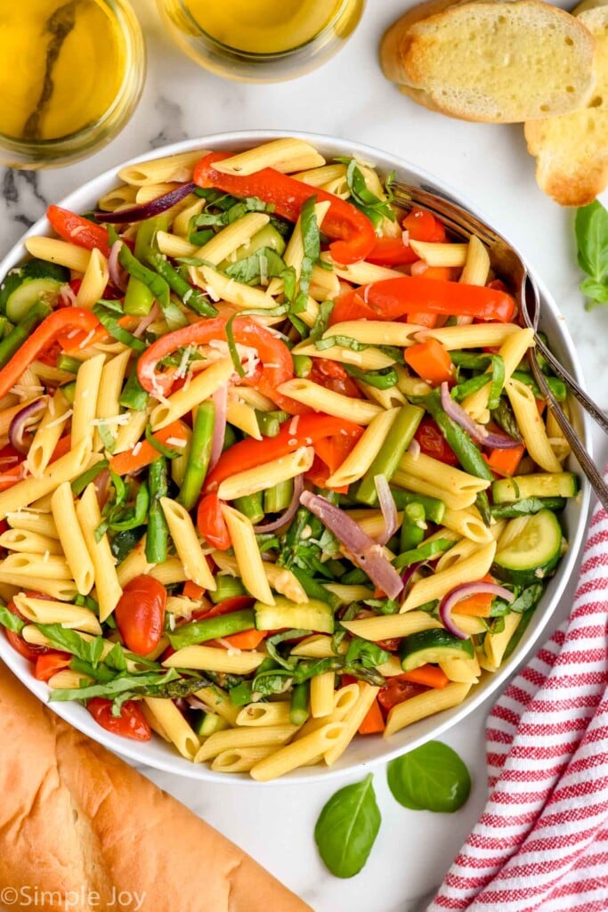 Overhead view of a bowl of Pasta Primavera garnished with shredded parmesan and basil, with forks. Bread, glasses of white wine beside.