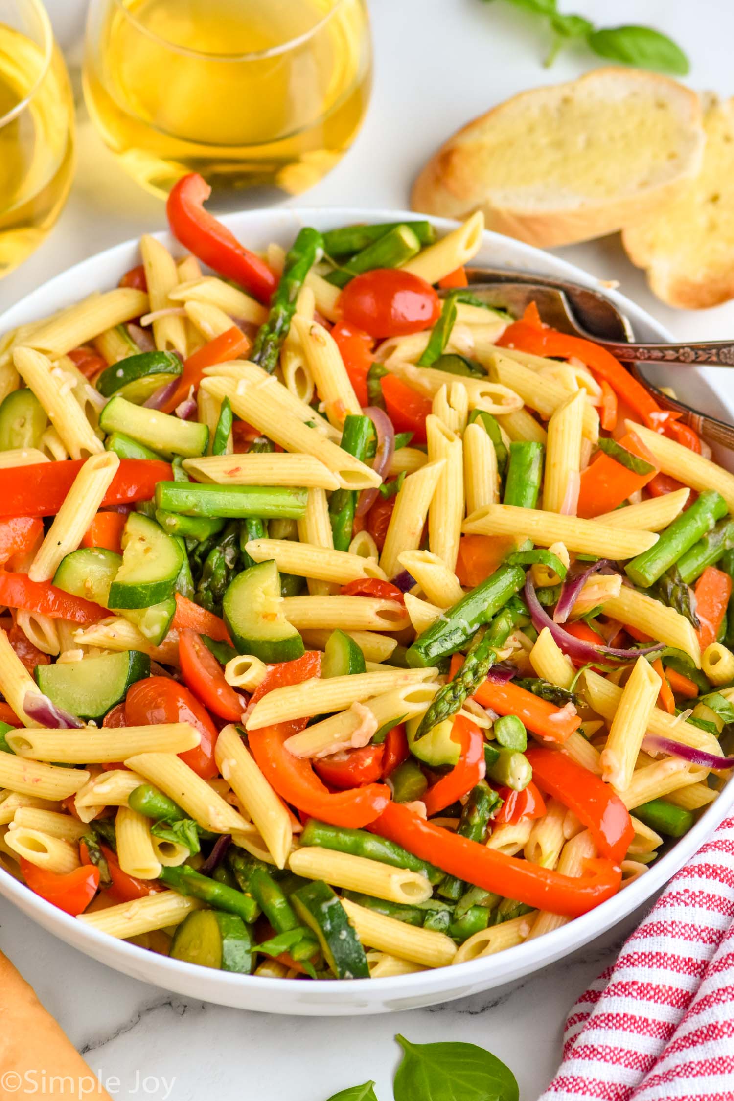 Bowl of Pasta Primavera with forks for serving. Bread and glasses of white wine beside.