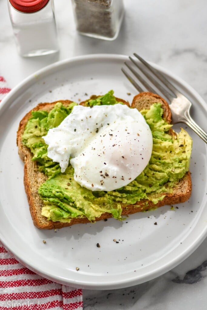 piece of toast on a plate topped with smashed avocado and poached egg topped with black pepper with fork sitting beside. Salt and pepper shakers sitting behind