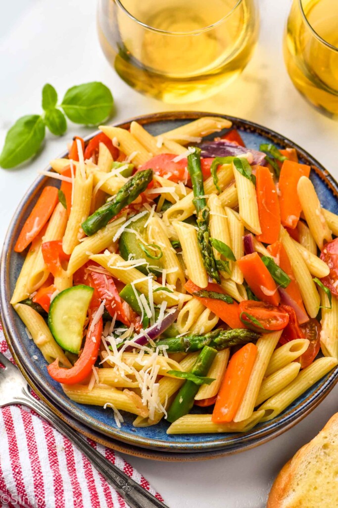Overhead view of a plate of Pasta Primavera garnished with shredded parmesan cheese. Fork and glasses of white wine beside.