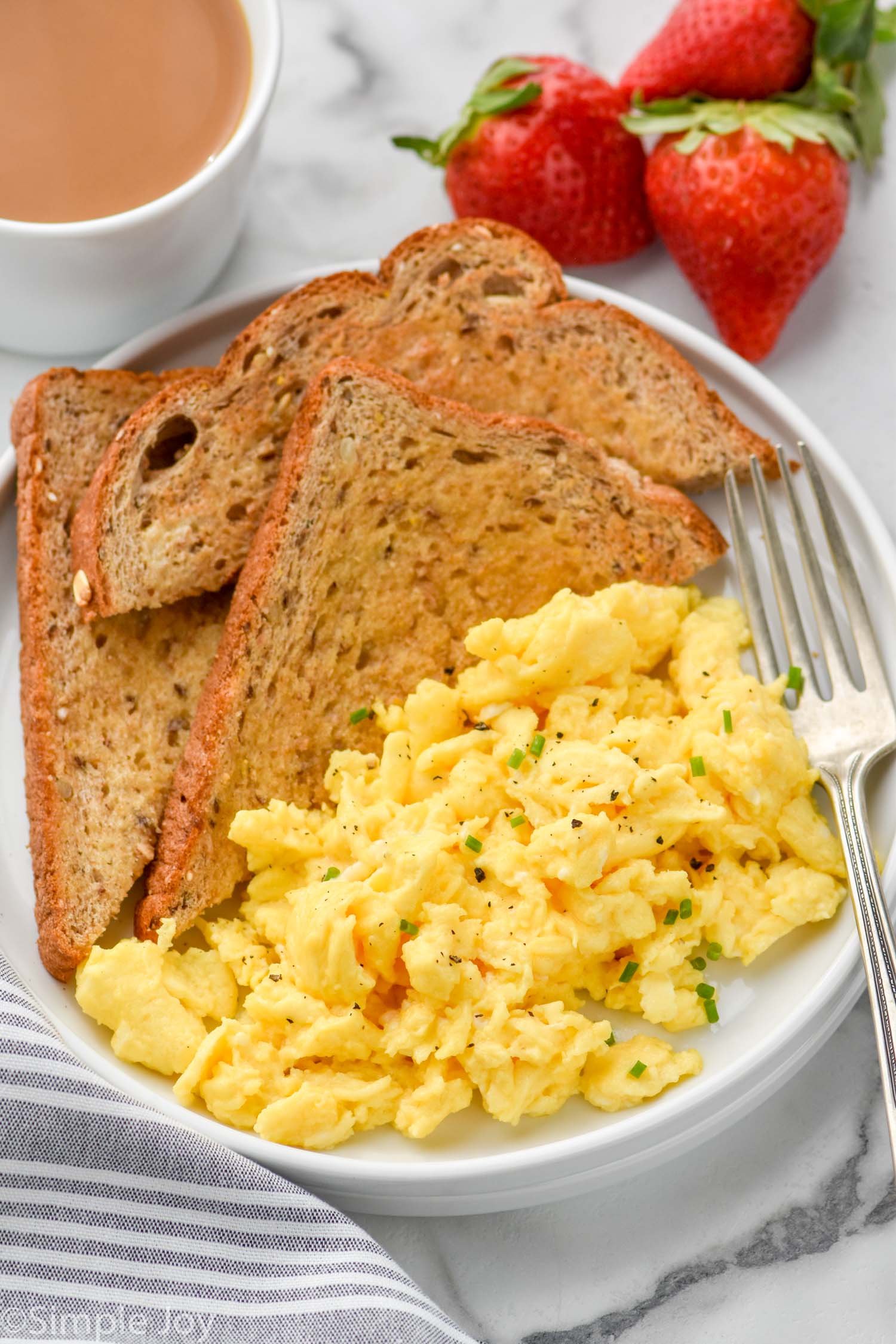 plate of Scrambled Eggs, pieces of toast, and a fork. Fresh strawberries and a mug of coffee sitting beside