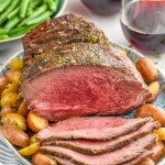 Platter with Sirloin Tip Roast partially sliced, with potatoes. Bowl of green beans and glasses of red wine beside. - 12
