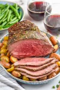 Platter with Sirloin Tip Roast partially sliced, with potatoes. Bowl of green beans and glasses of red wine beside.