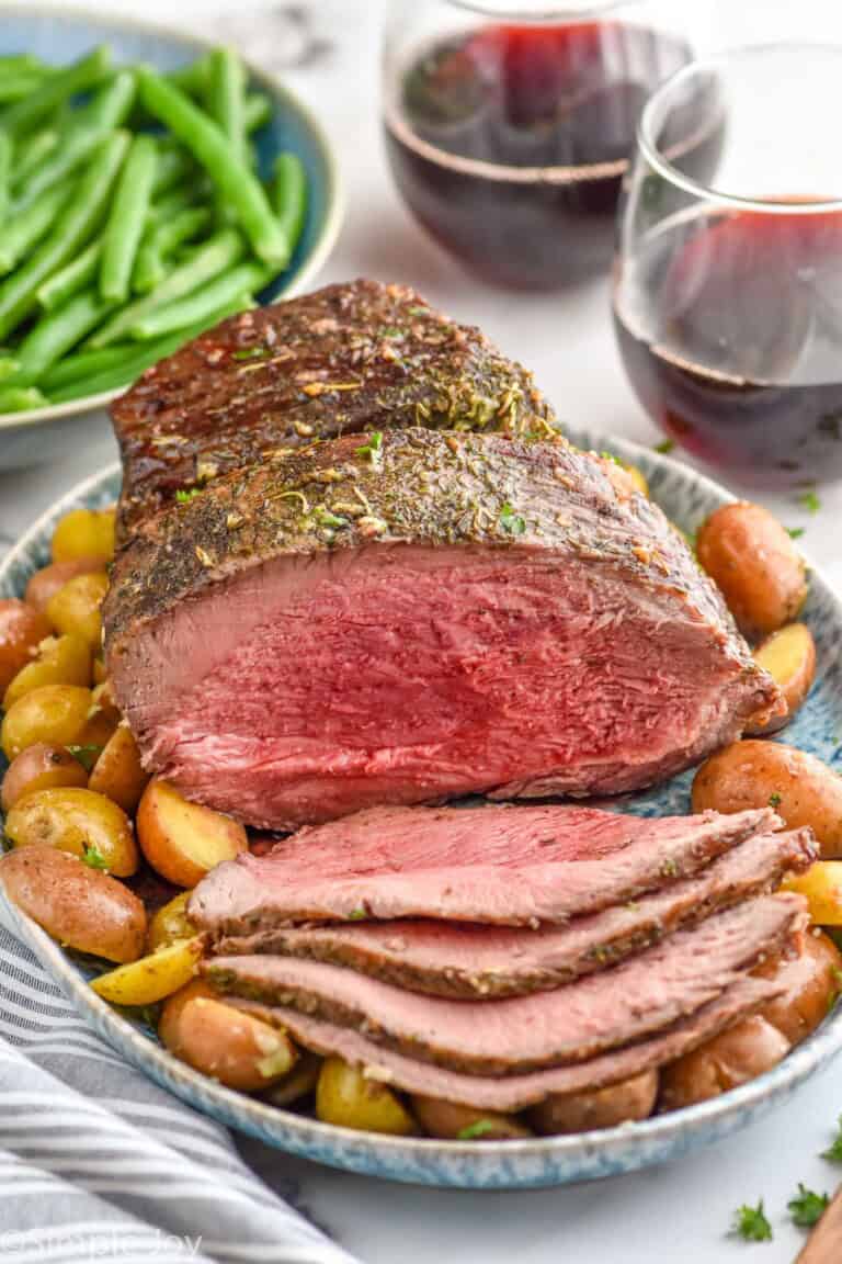 Platter with Sirloin Tip Roast partially sliced, with potatoes. Bowl of green beans and glasses of red wine beside.