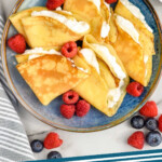 Pinterest graphic for Crepes. Image shows overhead of plate of crepes with whipped cream and fresh raspberries. Two mugs and coffee and fresh berries sitting beside. Says "crepe recipe simplejoy.com"