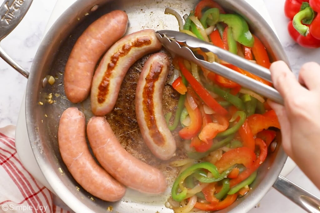 overhead of person's hand holding metal tongs in a skillet of diced bell peppers, yellow onion, and italian sausages to make sausage and peppers. Red bell pepper and green bell pepper sitting beside.