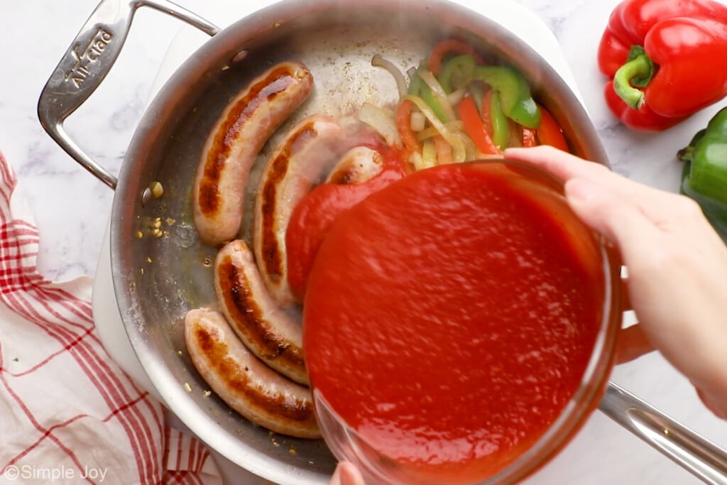 overhead of bowl of tomato sauce pouring into a skillet of diced peppers, onions, and sausages to make sausage and peppers.