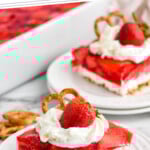 Pinterest graphic for Strawberry Pretzel Salad. Says "super easy Strawberry Pretzel Salad simplejoy.com" Image shows a piece of Strawberry Pretzel Salad topped with whipped cream and a strawberry on a plate. Pan and plate of Strawberry Pretzel Salad sitting in background