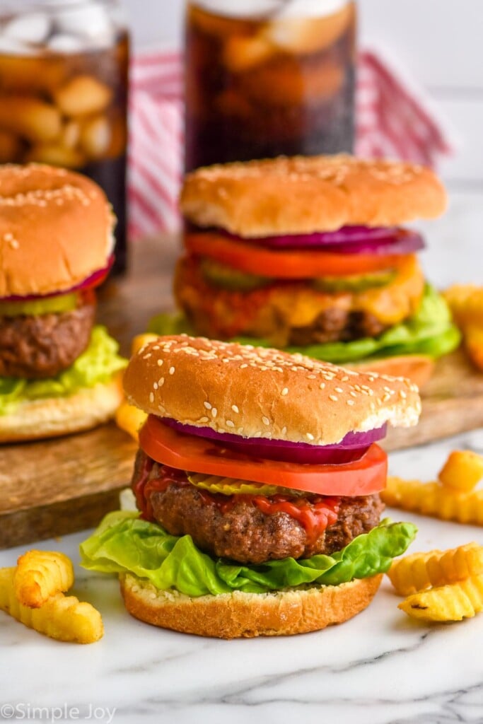 Grilled hamburger on a but with lettuce, pickles, tomato slice, red onion, and ketchup. Hamburgers sitting in background, french fries sitting beside.