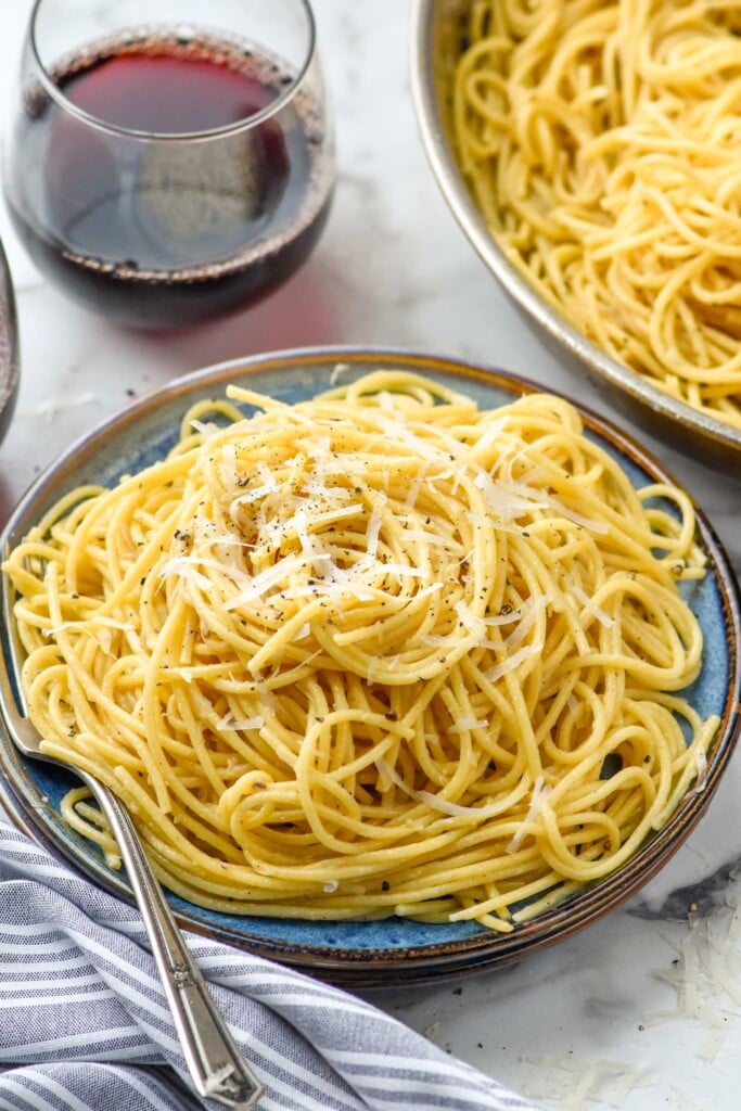 Plate of Cacio E Pepe topped with ground black pepper and grated cheese. Glass of red wine and skillet of Cacio E Pepe sitting in background.