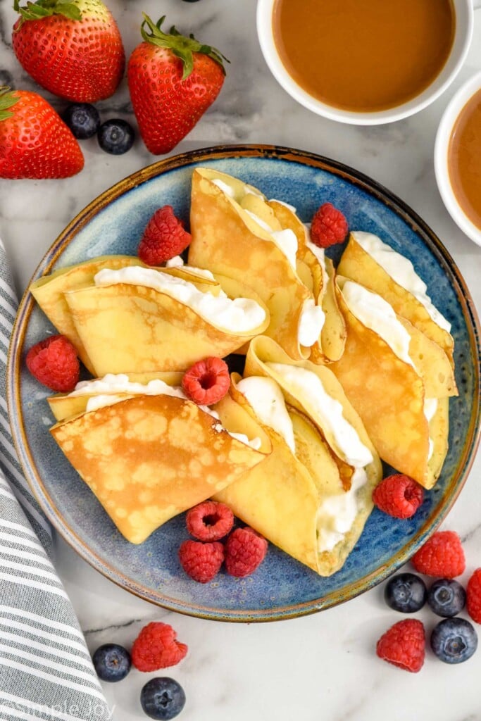 Overhead of plate of crepes stuffed with whipped cream and topped with fresh berries. two mugs of coffee and berries sitting beside plate.