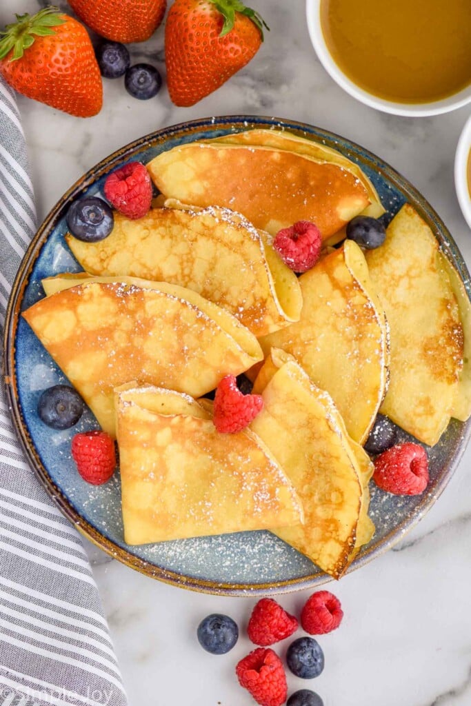 Overhead of plate of homemade crepes topped with powdered sugar and fresh fruit. Mug of coffee and more fresh berries sitting beside