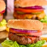 Grilled hamburger on a but with lettuce, pickles, tomato slice, red onion, and ketchup. Hamburgers sitting in background