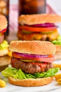 Grilled hamburger on a but with lettuce, pickles, tomato slice, red onion, and ketchup. Hamburgers sitting in background