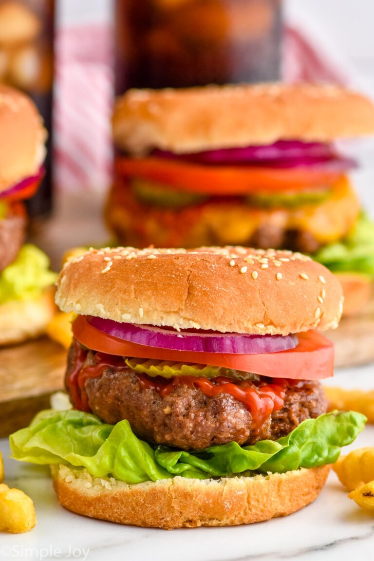 Grilled hamburger on a but with lettuce, pickles, tomato slice, red onion, and ketchup. Hamburgers sitting in background