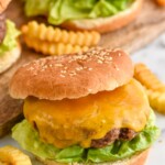Hamburger topped with cheese on a bun with lettuce. French fries and hamburgers sitting beside and behind